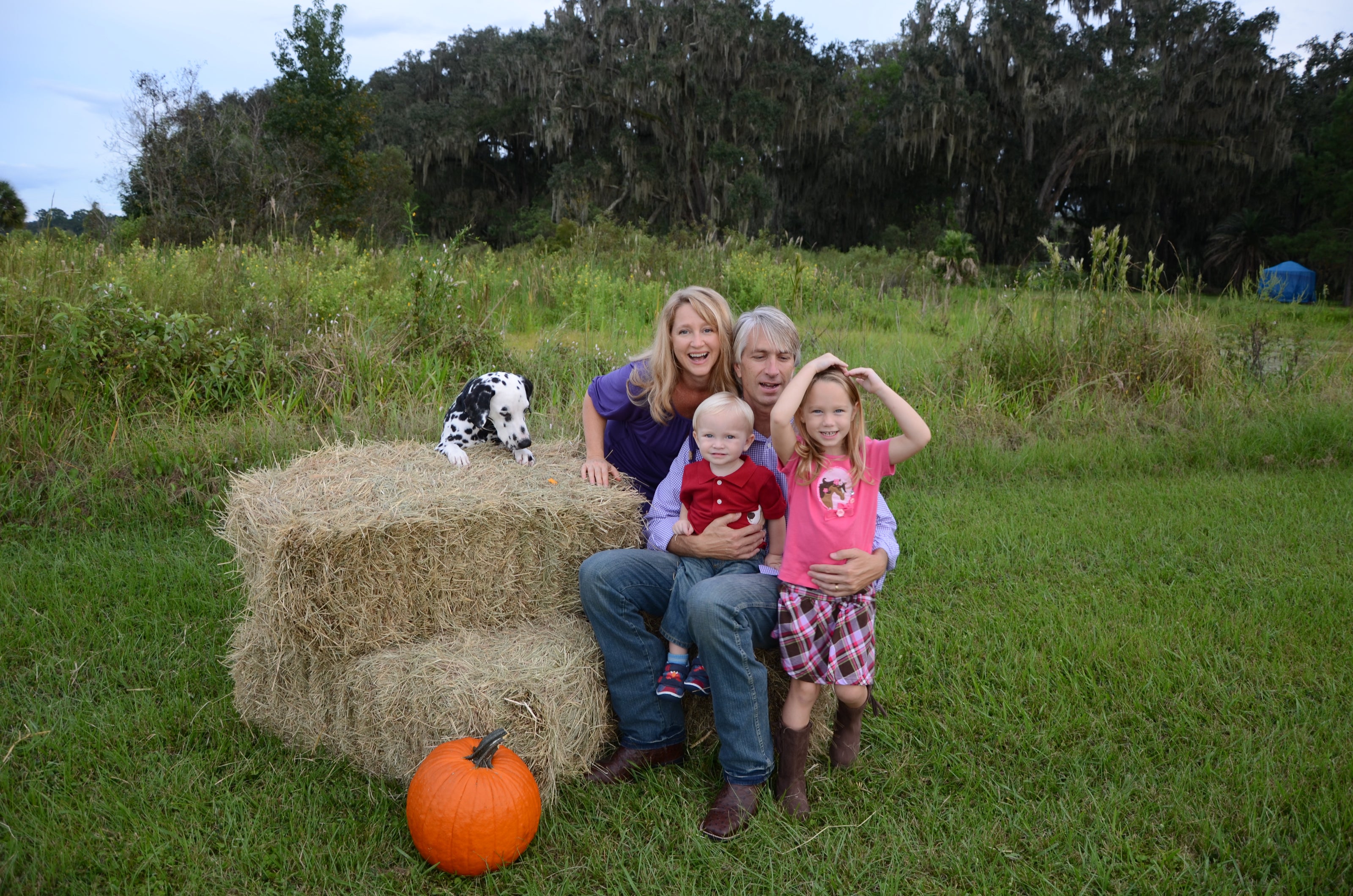 Family with two children and a dalmation dog sitting on hay bales in a grassy field with a pumpkin near St. Johns River.